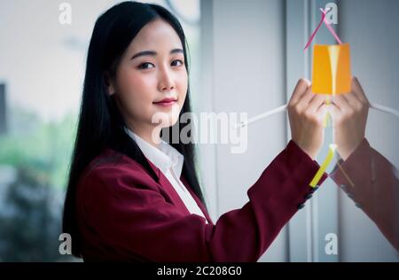 Porträt der jungen asiatischen Geschäftsfrau Schreiben klebrigen Papier Pad auf Glas in modernen Büro. Businessplan-Konzept. Stockfoto