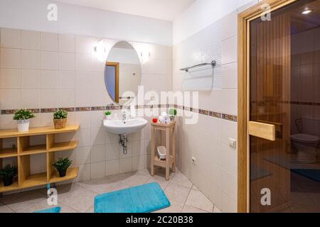 Modernes Interieur mit geräumigem Bad mit Sauna. Hellbeige Fliesen. Weißes Waschbecken mit Spiegel. Blumen auf den Holzregalen. Stockfoto