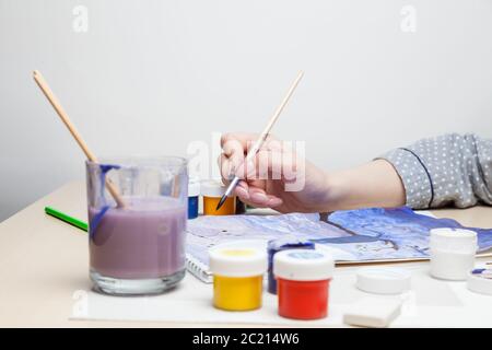 Weibliche Hand hält einen Pinsel und zeichnet Bild am Tisch in der Mitte von Gläsern mit Aquarellfarben und daneben ist ein Glas zum Spülen und Reinigen der b Stockfoto
