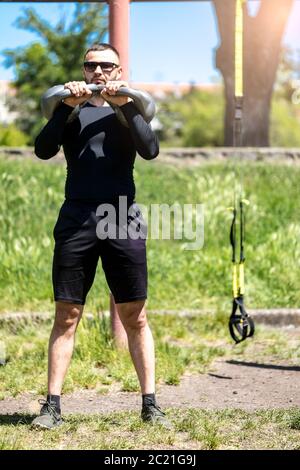 Starker junger Mann, der Schultern mit Kettle Bell Gewichten trainiert. Mann trainiert mit Kettlebell bei sonnigem Wetter. Stockfoto