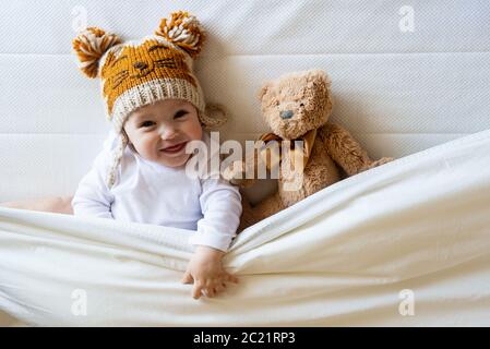 Happy Boy mit Wollmütze, der mit Teddybär unter der Decke auf dem Bett liegt Stockfoto