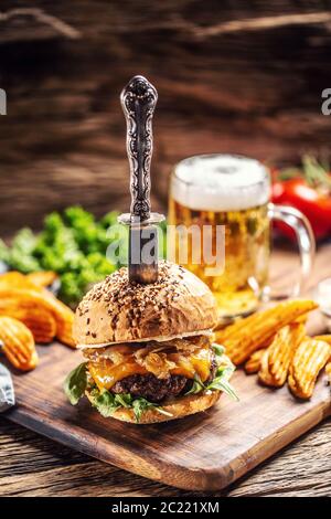 Rinderburger mit karamellisierten Zwiebeln und Rucola in rustikaler Holzumgebung, ein Bier und Kartoffelkeile an der Seite Stockfoto