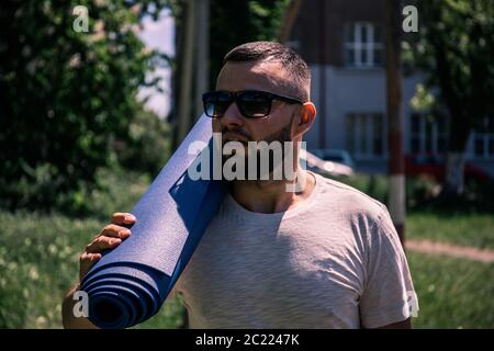 Junger bärtiger Sportler mit einer blauen Matte im Park. Junger Mann praktiziert Yoga Fitness-Übung im Freien. Stockfoto