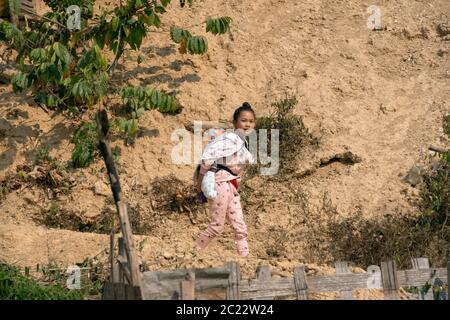 Ein junges, indigenes, ethnisches Lanten-Mädchen, das in ihrem Dorf in der Provinz Luang Namtha im Norden Laos ein Baby trägt. Stockfoto