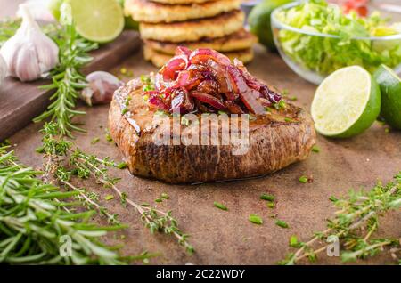 Leckeres Rindersteak mit Pfeffer, Knoblauch und karamellisierten Zwiebeln, Kartoffelpuffer hinter Stockfoto