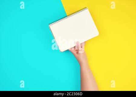 Woman's Hand hält offene Spirale Notebook mit leeren weißen Blätter auf einem gelb-blauen Hintergrund