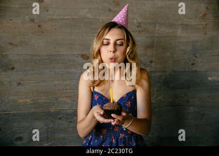 Nette junge Frau ausblasen Kerze zum Geburtstag chocolate Cupcake. Festliche Hut, lockiges Haar. Urlaub Konzept. Stockfoto