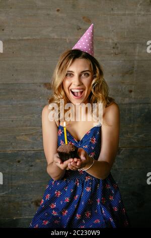 Glücklich attraktives Mädchen hält einen chocolate Cupcake mit Kerze, Ihren Geburtstag feiern. In blauem Kleid gekleidet, in festlicher Hut. Urlaub Konzept. Stockfoto