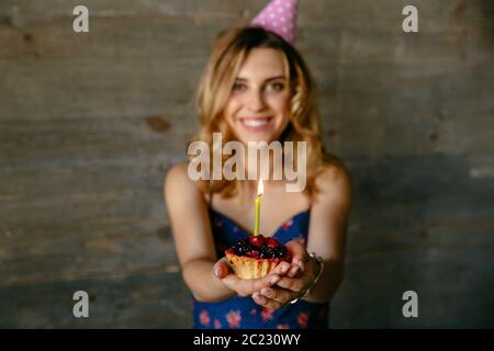 Glücklich lächelnde junge Frau feiert ihren Geburtstag, hält eine Frucht Cupcake mit Kerze. In festlichen Hut. Urlaub Emotionen. Stockfoto