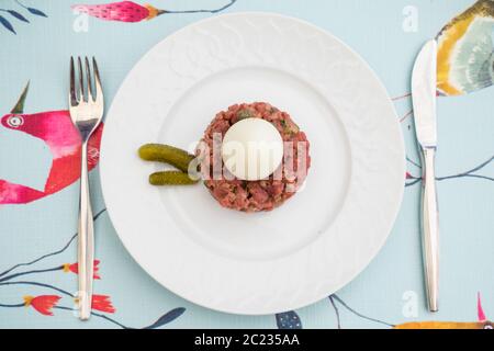 Rohes Steak tatar mit gekochtem Ei und Gurken auf einem weißen Teller mit Besteck auf einem dekorativen Tisch Stockfoto