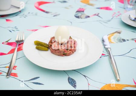 Rohes Steak tatar mit gekochtem Ei und Gurken auf einem weißen Teller mit Besteck auf einem dekorativen Tisch Stockfoto
