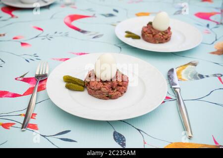 Rohes Steak tatar mit gekochtem Ei und Gurken auf einem weißen Teller mit Besteck auf einem dekorativen Tisch Stockfoto