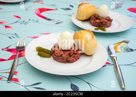 Rohes Steak tatar mit gekochtem Ei und Gurken auf einem weißen Teller mit Besteck auf einem dekorativen Tisch Stockfoto