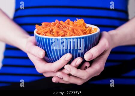 Weibliche Hände, die eine Schüssel mit orangefarbenen Linsenfusilli-Pasta auf einem blau gestreiften Hintergrund halten. Diät- und glutenfreies Konzept Stockfoto