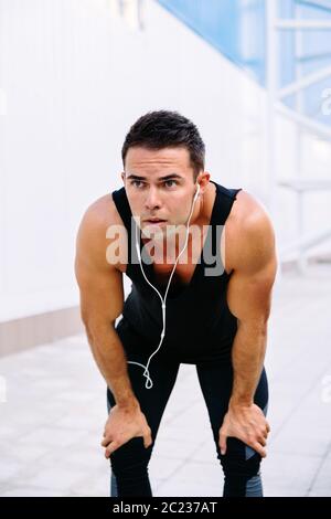 Portrait von müde Sportler, die nach dem Ausführen, halten die Hände auf seine Knie, hören die Musik der Ohrhörer, mit schwarzen Sportswear. Stockfoto