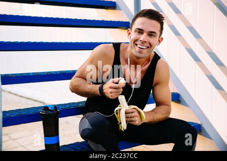 Lächelnd sportlicher Kerl der Ohrhörer, eine Banane essen, nach dem Training, sitzen auf den Treppen und an der Kamera, im Freien. Stockfoto