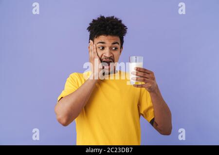 Foto von schockierten afroamerikanischen Mann hält Glas mit Milch isoliert über lila Hintergrund Stockfoto