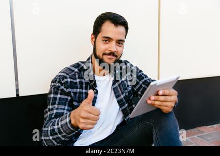 Attraktive bärtiger Mann mit einem Daumen hoch, von der Kamera suchen, während Sie einen digitalen Tablet und sitzen in der Nähe der Wand, im Freien. Stockfoto