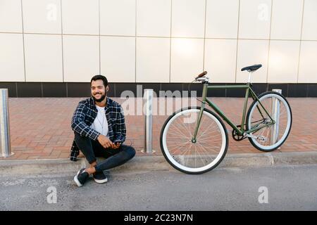 Outdoor Foto von stilvollen attraktiver Mann in der Nähe der Bike sitzen, Musik hören und der Ohrhörer, Genießen der Freizeit draußen. Stockfoto