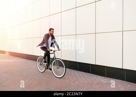 Volle Länge Foto von lächelnden Jungen bärtigen Mann mit dem Fahrrad auf der Strasse der Stadt. Im karierten Hemd, T-Shirt und Jeans. Erholung Konzept. Stockfoto