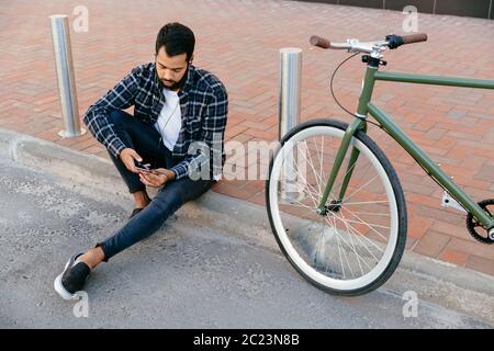 Blick von oben auf die Jungen bärtigen Mann im Kopfhörer, mit seinem Mobiltelefon, ein Video ansehen, während in der Nähe das Fahrrad draußen sitzen. Stockfoto