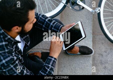 Nahaufnahme der Jungen stilvoll mit einem Tablet Mann, Durchsuchen von Websites beim Sitzen auf die Schritte in der Nähe der Bike, im Freien. Stockfoto