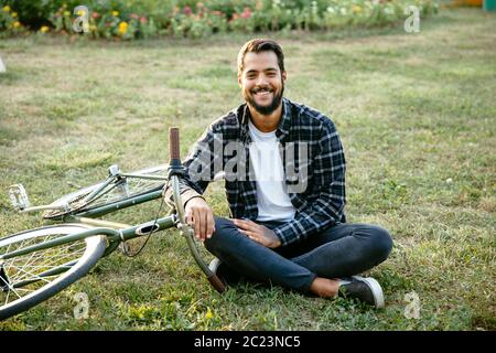 Lächelnd bärtiger Mann im Park entspannen, sitzen auf Gras in der Nähe des Fahrrades, auf Kamera. Freizeitaktivitäten Konzept. Stockfoto