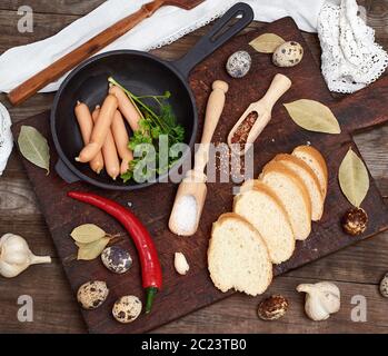 Würstchen in einem schwarzen runden gusseisernen Pfanne und raw Wachteleier auf einem grauen Holz- Tabelle, Ansicht von oben Stockfoto