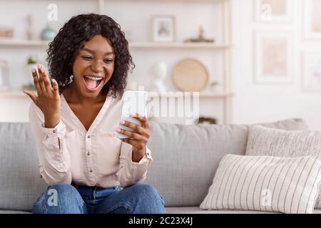 Überglücklich schwarzes Mädchen feiert Erfolg mit Smartphone zu Hause, erhielt gute Nachrichten Stockfoto
