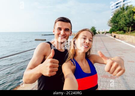 Lächelnd Sportlerin und Sportler in Sportkleidung mit einem Daumen hoch, während eine selfie während des Trainings auf dem Kai, in der Nähe des Meeres. Stockfoto