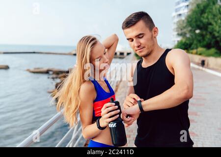 Muskulöse Mann und Fitness Mädchen überprüfung auf Uhren nach dem Lauftraining auf dem Kai, in der Nähe des Meeres. Sport Konzept. Stockfoto