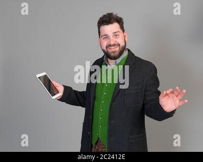 Jung und cool Österreichischer Mann mit schwarzem Haar und Bart in der traditionellen Tracht steht vor einem grauen Hintergrund und arbeitet auf einem Tablet Stockfoto