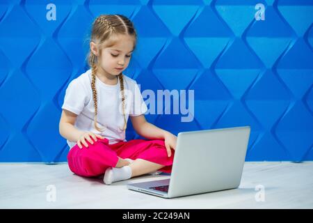 7 Jahre altes Mädchen in einem weißen T-Shirt sitzt mit einem Laptop auf dem Boden und drückt die Tasten Stockfoto