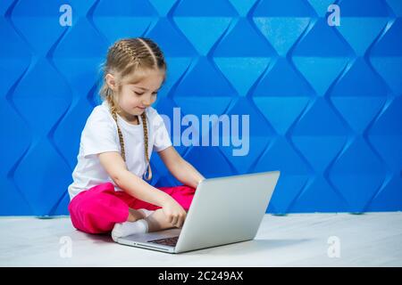 7 Jahre altes Mädchen in einem weißen T-Shirt sitzt mit einem Laptop auf dem Boden und drückt die Tasten Stockfoto