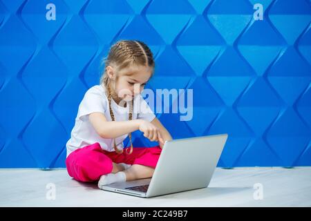 7 Jahre altes Mädchen in einem weißen T-Shirt sitzt mit einem Laptop auf dem Boden und drückt die Tasten Stockfoto