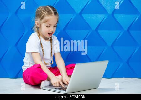 7 Jahre altes Mädchen in einem weißen T-Shirt sitzt mit einem Laptop auf dem Boden und drückt die Tasten Stockfoto