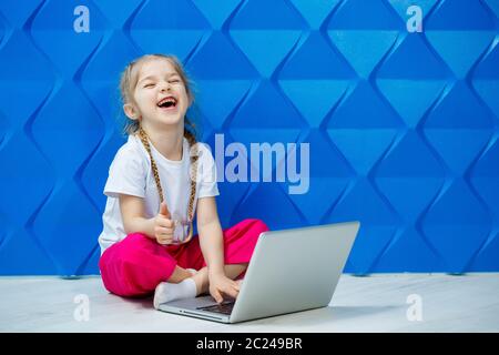 7 Jahre altes Mädchen in einem weißen T-Shirt sitzt mit einem Laptop auf dem Boden und drückt die Tasten Stockfoto