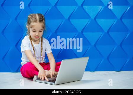 7 Jahre altes Mädchen in einem weißen T-Shirt sitzt mit einem Laptop auf dem Boden und drückt die Tasten Stockfoto