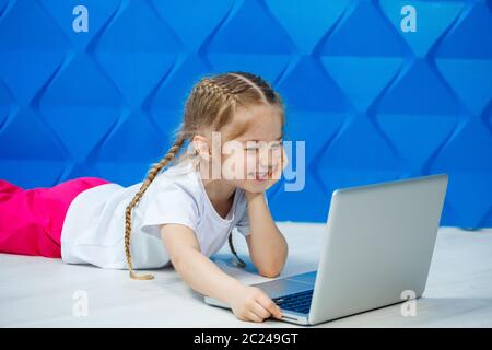 7 Jahre altes Mädchen in einem weißen T-Shirt sitzt mit einem Laptop auf dem Boden und drückt die Tasten Stockfoto