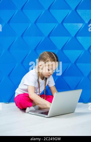 7 Jahre altes Mädchen in einem weißen T-Shirt sitzt mit einem Laptop auf dem Boden und drückt die Tasten Stockfoto