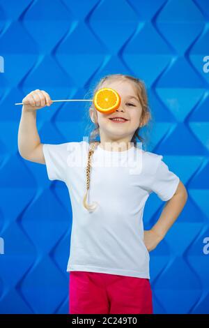 Ein kleines Mädchen im T-Shirt lächelt und hält zwei Hälften oranger Früchte vor ihren Augen auf blauem Hintergrund. Konzept: Der Kampf gegen das Frühjahr AV Stockfoto