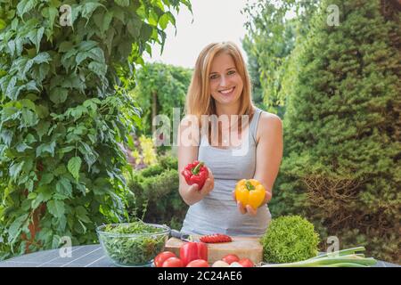 Lächelnd attraktive junge Frau ist mit roten und gelben Paprika im Freien. Andere frische Gemüse liegen auf dem Tisch. Stockfoto