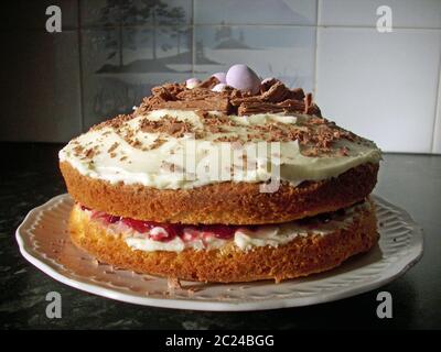 Hausgemachte Osterkuchen aus Biskuit mit Marmelade Füllung und Schokolade Flocke und Osterei Topping. Auf einer weißen Platte. Von der Seite beleuchtet. Hintergrund von Stockfoto