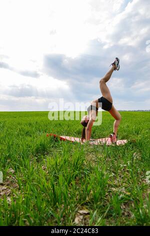 Eine junge Frau in einem schwarzen Top und Shorts führt einen Handstand. Ein Model steht auf ihren Händen, macht Gymnastik Splits gegen den blauen Himmel. Gesund am lebendigsten Stockfoto