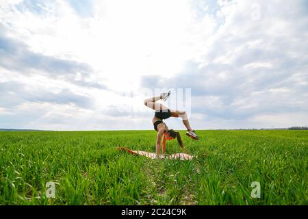 Schöne Mädchen Modell auf grünem Gras tun Yoga. Eine schöne junge Frau auf einem grünen Rasen führt akrobatische Elemente. Flexible Turnerin in schwarz tut ein han Stockfoto