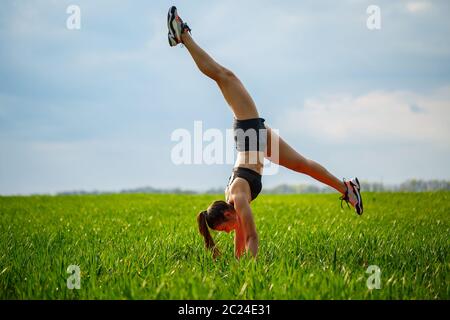 Schöne Mädchen Modell auf grünem Gras tun Yoga. Eine schöne junge Frau auf einem grünen Rasen führt akrobatische Elemente. Flexible Turnerin in schwarz tut ein han Stockfoto