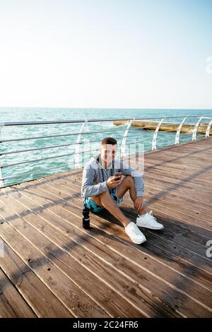 Lächelnd gutaussehenden Mann im Kopfhörer, Musik hören, telefonieren, ruht auf Pier, in der Nähe des Ozeans, Sportkleidung zu tragen. Stockfoto