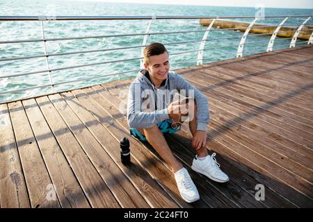 Glückliche junge Sportler Mann im Kopfhörer, Musik hören, telefonieren, im Internet surfen, chatten, während er auf Pier, in der Nähe des Meeres, das Tragen von spo Stockfoto