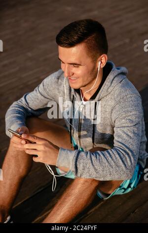 Freudige gutaussehenden Sportler der Ohrhörer, Musik hören, während Sie ein Mobiltelefon, Ausruhen nach Training, sitzen auf dem Boden, das Tragen sport jac Stockfoto