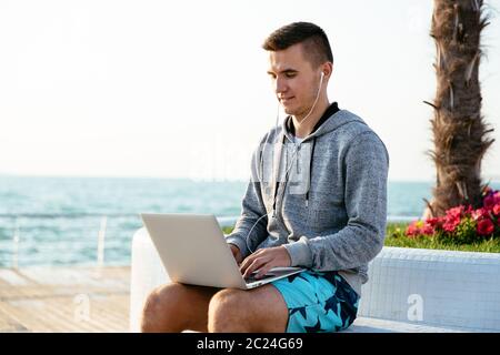 Junger hübscher Kerl in Kopfhörer Arbeiten am Laptop, beim Sitzen auf der Bank, Sportkleidung zu tragen. In der Nähe des Meeres. Stockfoto